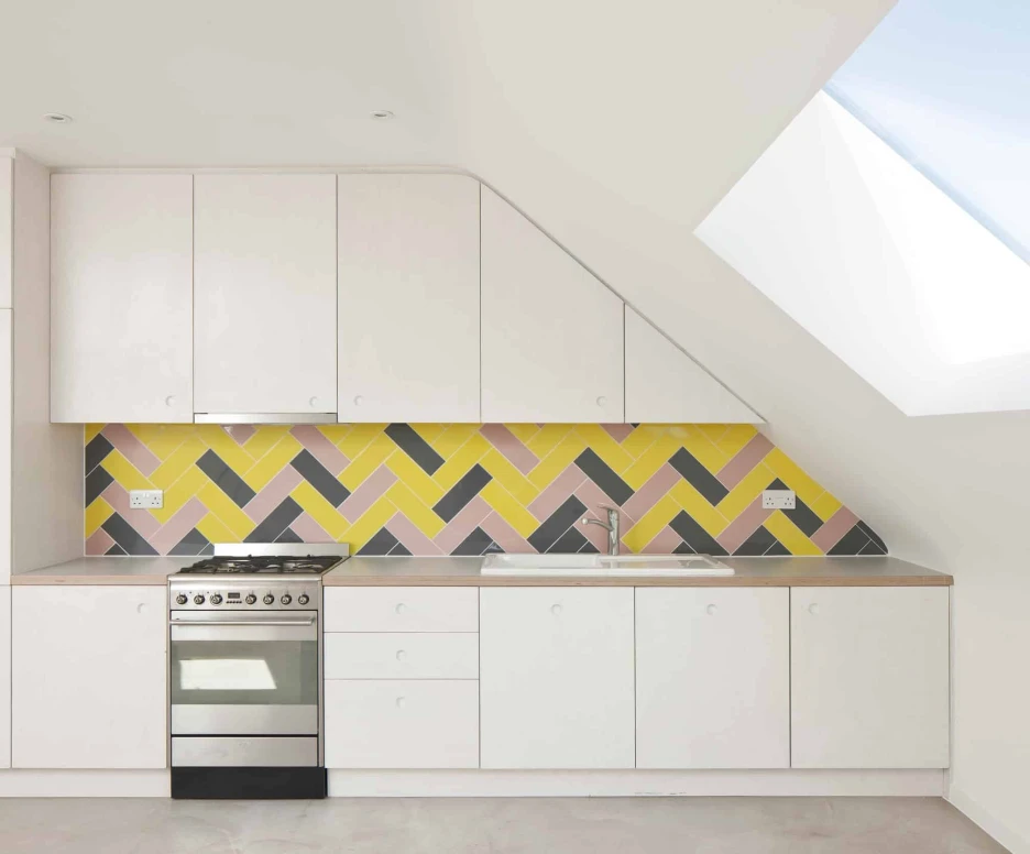 Cuisine moderne avec des armoires blanches, une hotte en inox, et un carrelage mural coloré en motifs zigzag jaune, rose et gris. Un puits de lumière illumine l'espace.