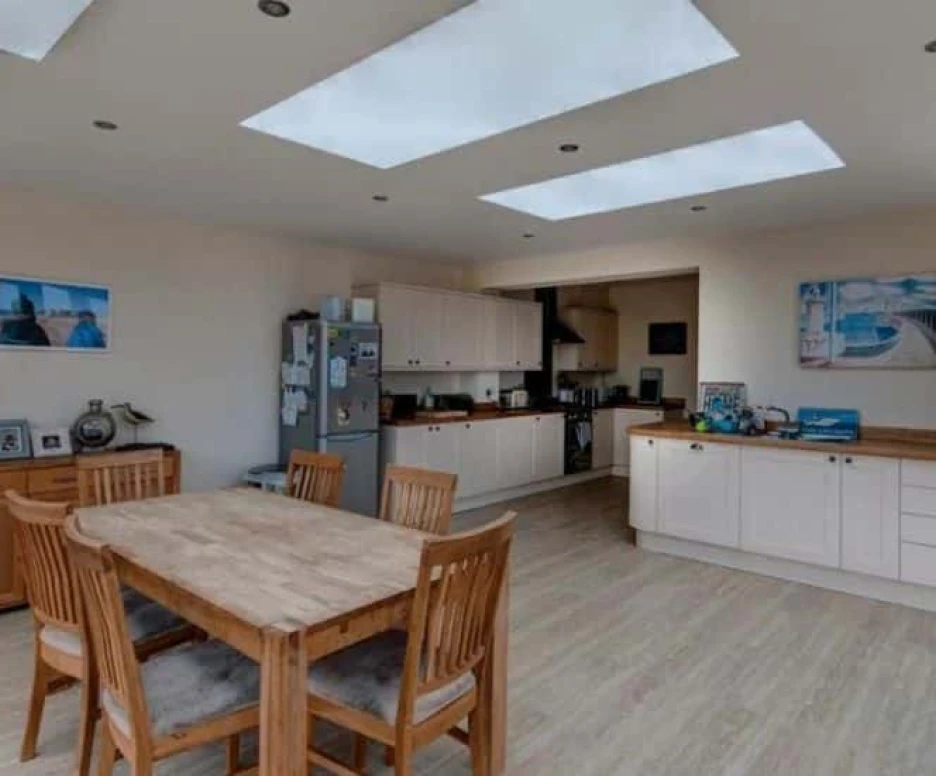 A modern kitchen and dining area featuring a wooden table with chairs, a potted plant, and large skylights. Cabinets and appliances are neatly arranged, with light-colored walls and a warm, inviting atmosphere.