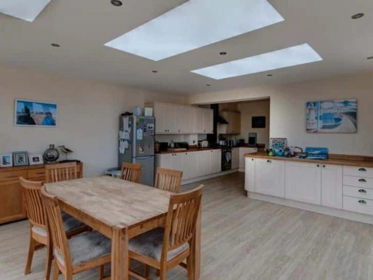 A modern kitchen and dining area featuring a wooden table with chairs, a potted plant, and large skylights. Cabinets and appliances are neatly arranged, with light-colored walls and a warm, inviting atmosphere.