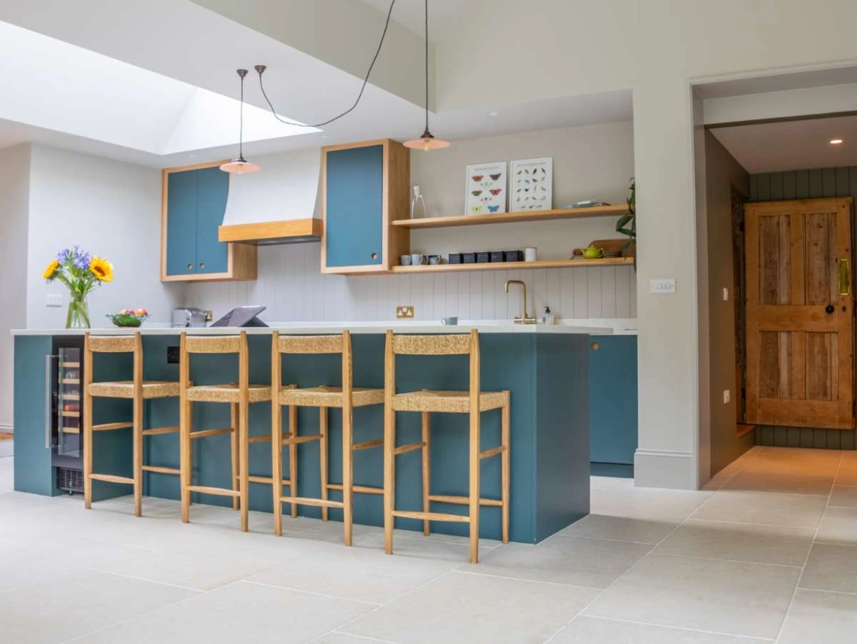 A modern kitchen with a large island featuring seating for four. Blue cabinetry complements light-colored walls and flooring. A door with wooden textures is visible in the background, and a vase with sunflowers adds a touch of color. Natural light floods the space from a skylight.