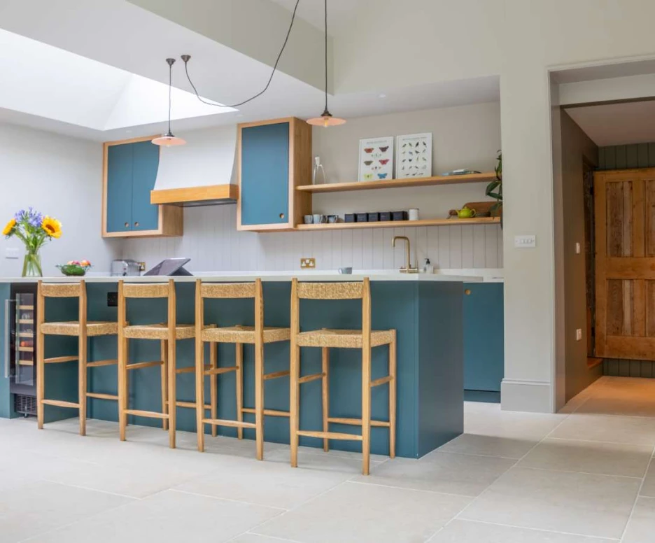 A modern kitchen with a large island featuring seating for four. Blue cabinetry complements light-colored walls and flooring. A door with wooden textures is visible in the background, and a vase with sunflowers adds a touch of color. Natural light floods the space from a skylight.