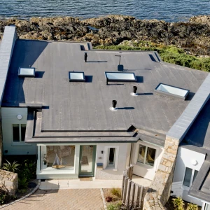 A modern coastal home with a flat roof and multiple skylights, situated near rocky shorelines and water. The exterior features a mix of stone and wood, with landscaped greenery surrounding the property.