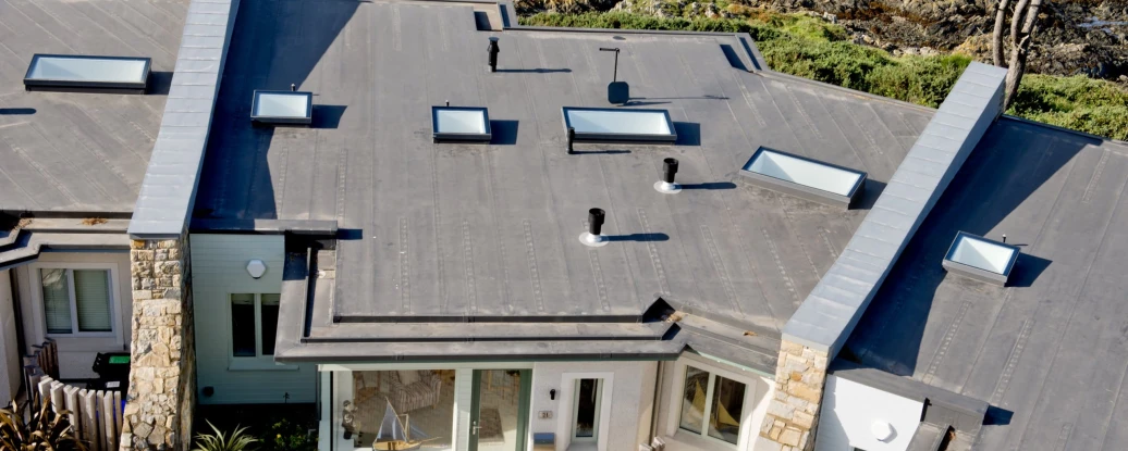 A modern coastal home with a flat roof and multiple skylights, situated near rocky shorelines and water. The exterior features a mix of stone and wood, with landscaped greenery surrounding the property.