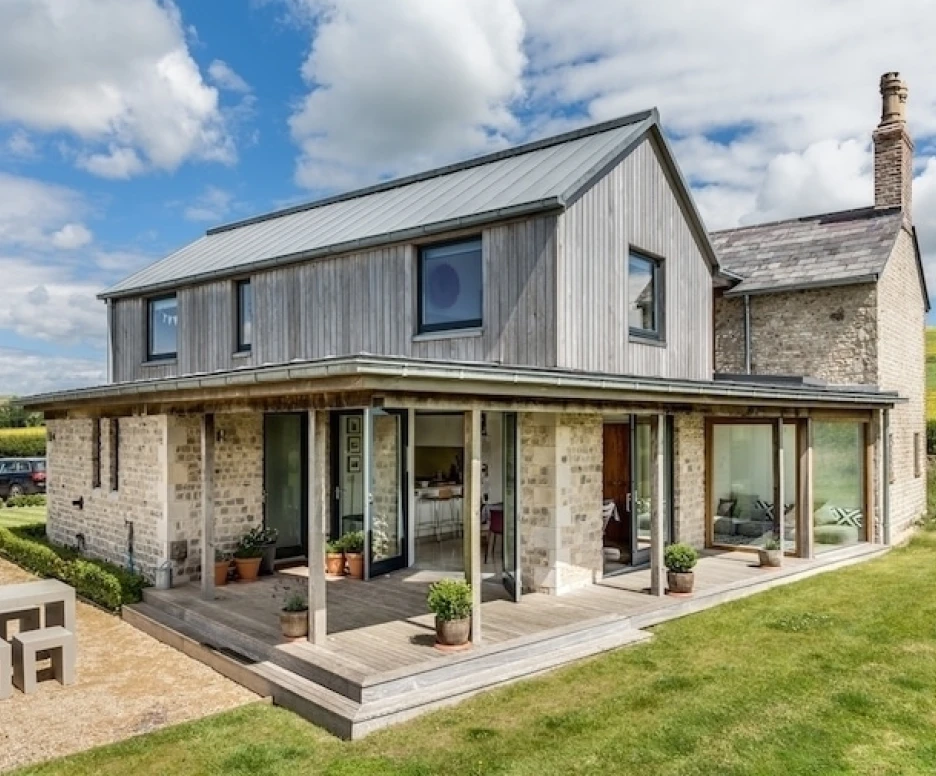 A modern two-story house featuring a wooden facade and large glass windows. The front porch is surrounded by potted plants, with a gravel driveway leading to the entrance. Lush green fields can be seen in the background under a partly cloudy sky.