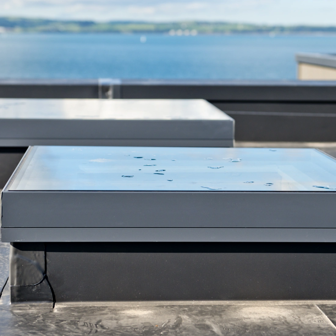 Zwei quadratische Dachfenster mit gläsernen Oberflächen stehen auf einem flachen, schwarzen Dach vor einem blauen Wasserhorizont.