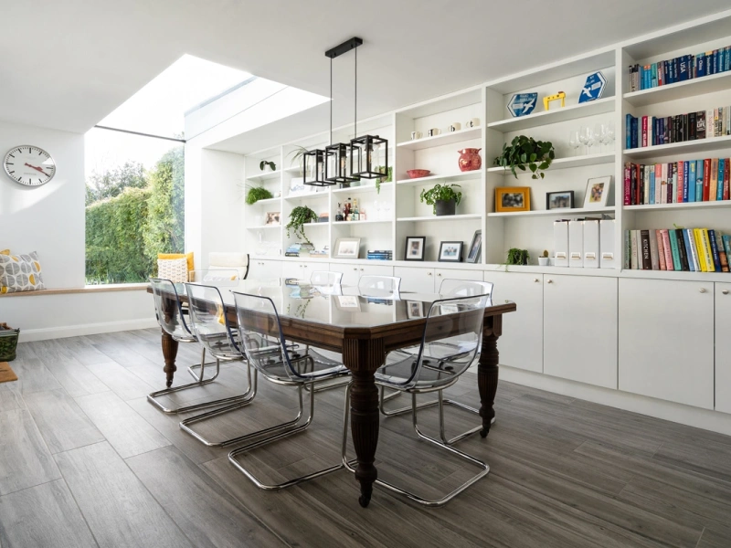 A modern kitchen and dining area features a long wooden table surrounded by transparent chairs. A large skylight above illuminates the space, highlighting built-in shelves filled with plants and colorful books. A cozy window seat is visible in the background.