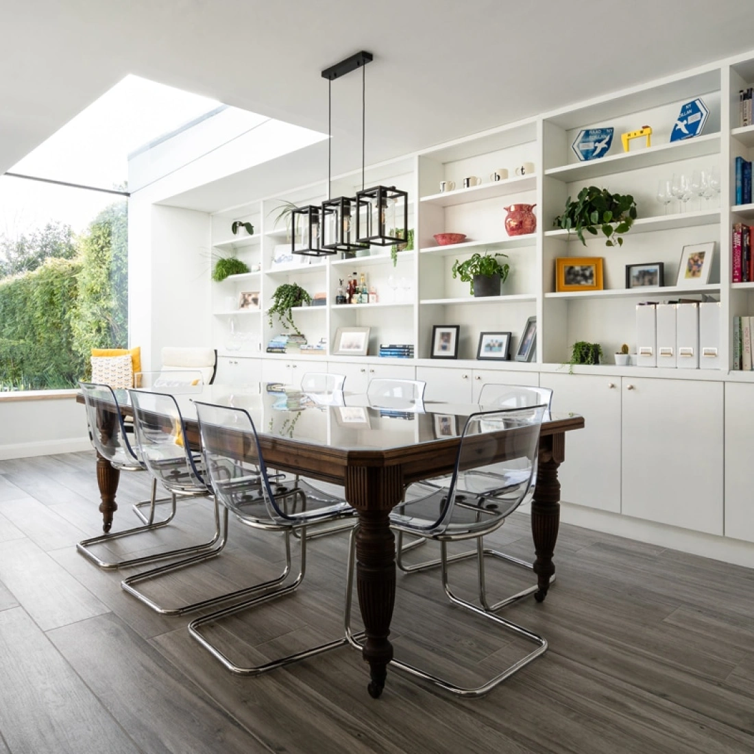 A modern dining area featuring a wooden table with transparent chairs. A large window illuminates the space, while shelves filled with books and decorative items line the walls. A cozy seating nook with yellow cushions is visible in the background.