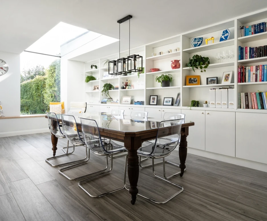 A modern dining area featuring a wooden table with transparent chairs. A large window illuminates the space, while shelves filled with books and decorative items line the walls. A cozy seating nook with yellow cushions is visible in the background.
