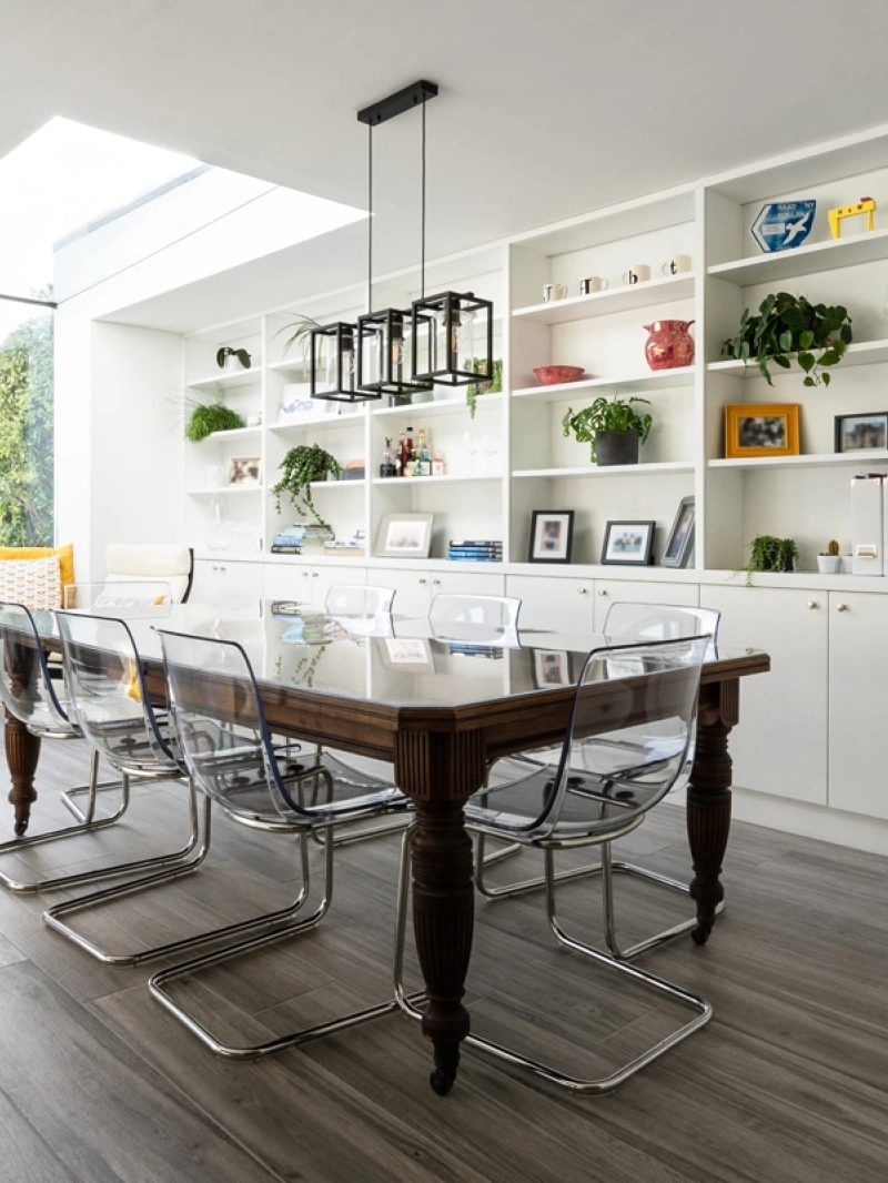 A modern dining area featuring a wooden table with transparent chairs. A large window illuminates the space, while shelves filled with books and decorative items line the walls. A cozy seating nook with yellow cushions is visible in the background.