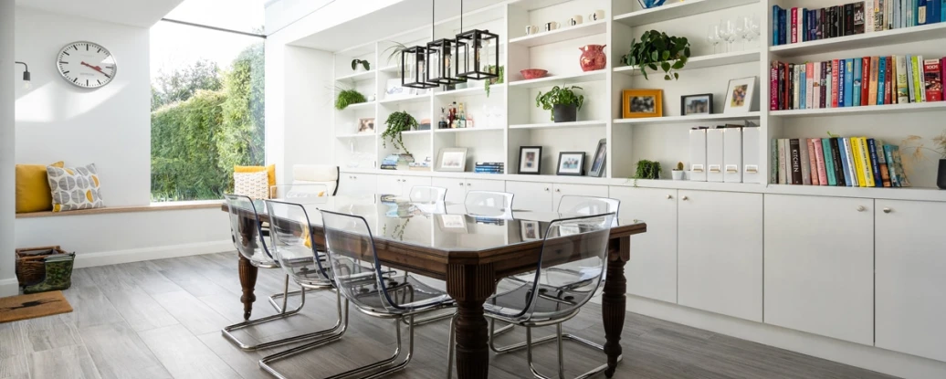 A modern dining area featuring a wooden table with transparent chairs. A large window illuminates the space, while shelves filled with books and decorative items line the walls. A cozy seating nook with yellow cushions is visible in the background.