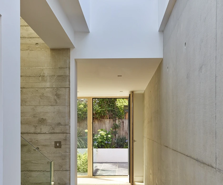 A modern hallway with smooth concrete walls, a wooden floor, and a glass railing. Light enters from an open door at the end, leading to a garden. The space features clean lines and a minimalist design.