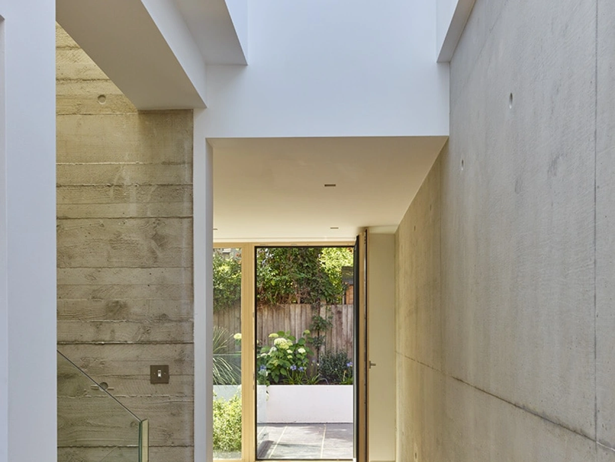 A modern hallway with smooth concrete walls, a wooden floor, and a glass railing. Light enters from an open door at the end, leading to a garden. The space features clean lines and a minimalist design.