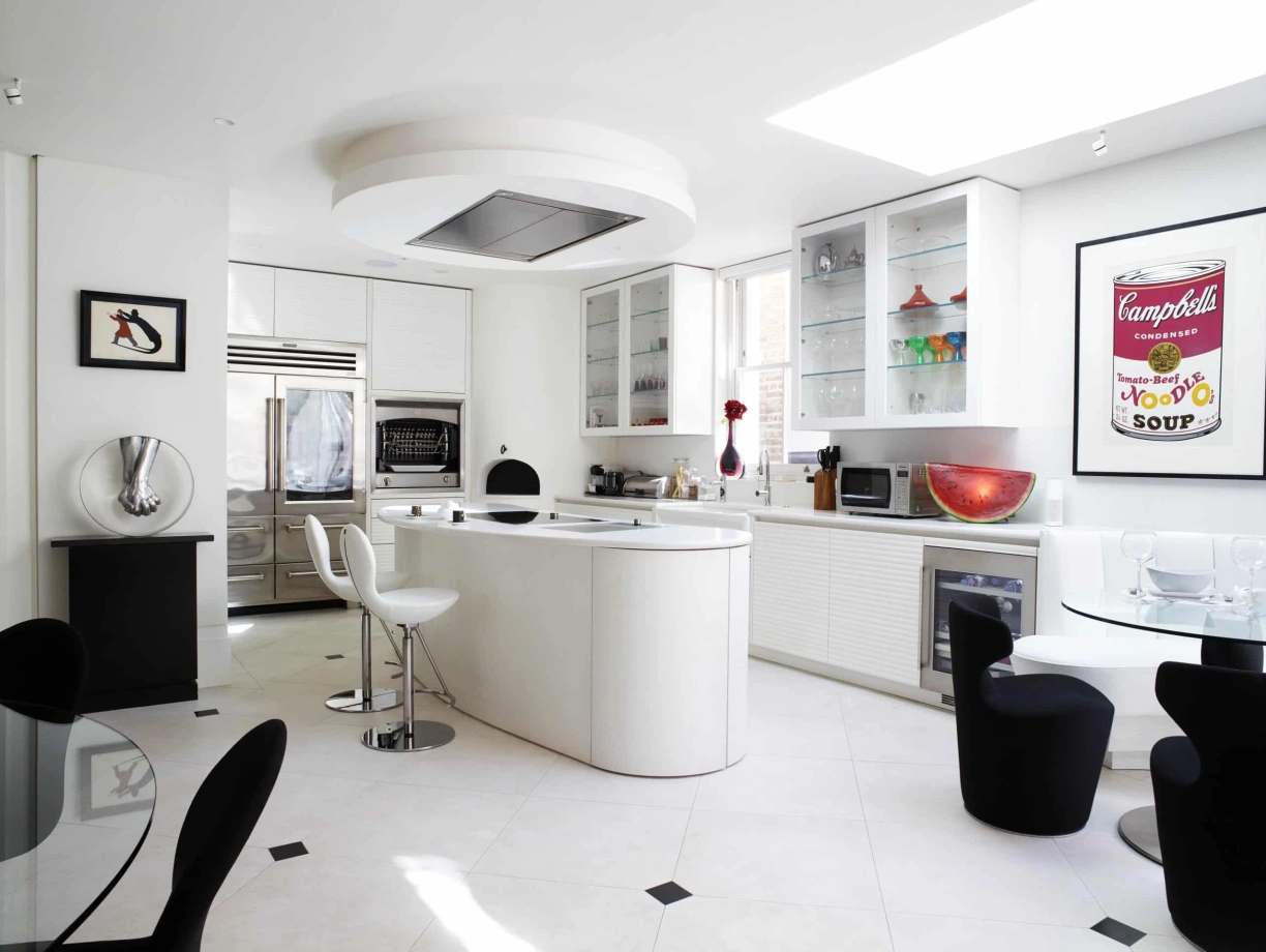 A modern kitchen featuring sleek white cabinetry, a large island with bar stools, and a stylish dining area. Bright natural light streams in through large windows, highlighting decorative glassware and a Campbell's Soup poster on the wall.