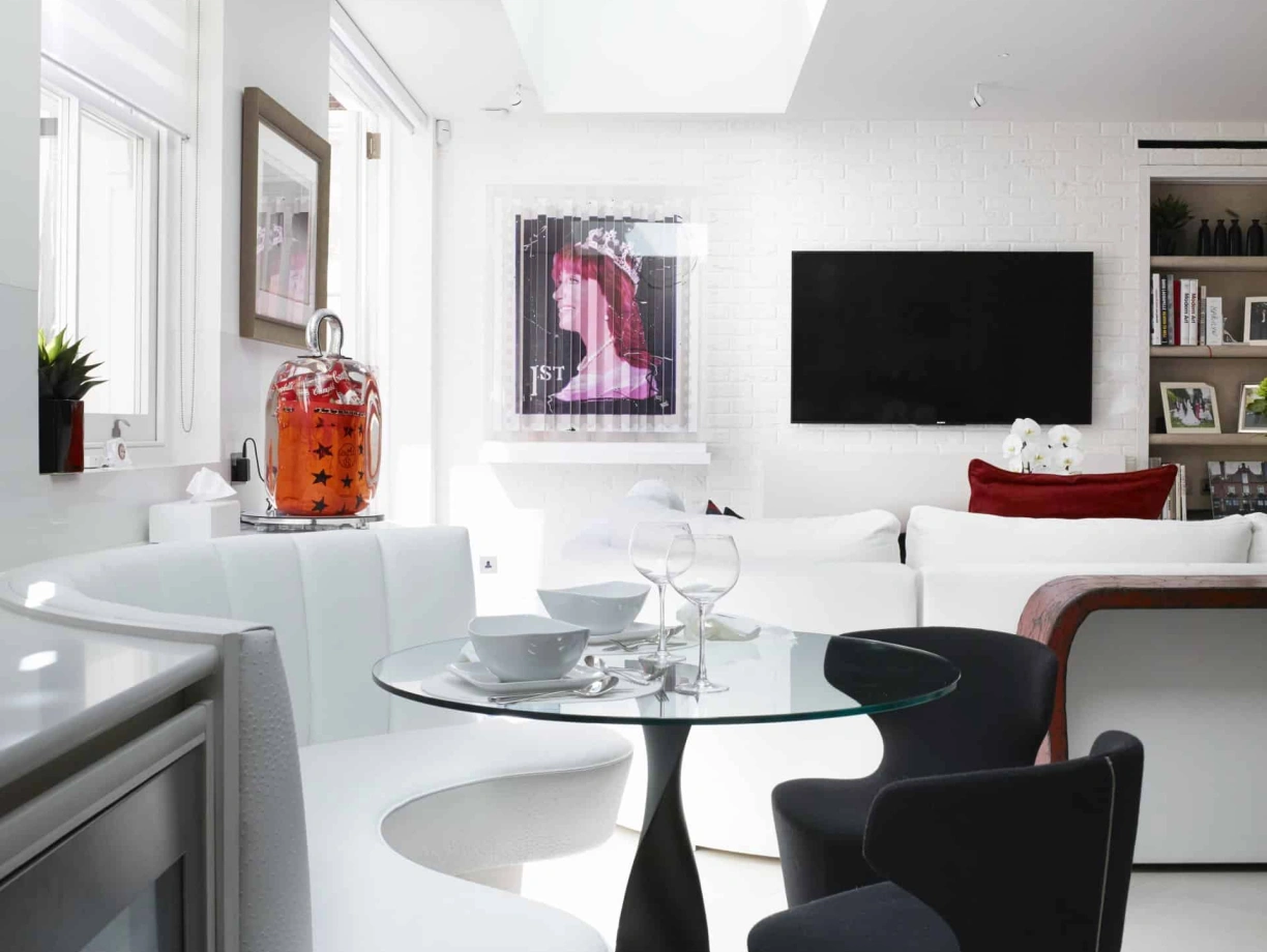A modern dining area featuring a round glass table with a sleek black chair and a curved white bench. The room has white walls and large skylights, creating a bright atmosphere. A black TV is mounted on the wall, and artwork is displayed nearby.