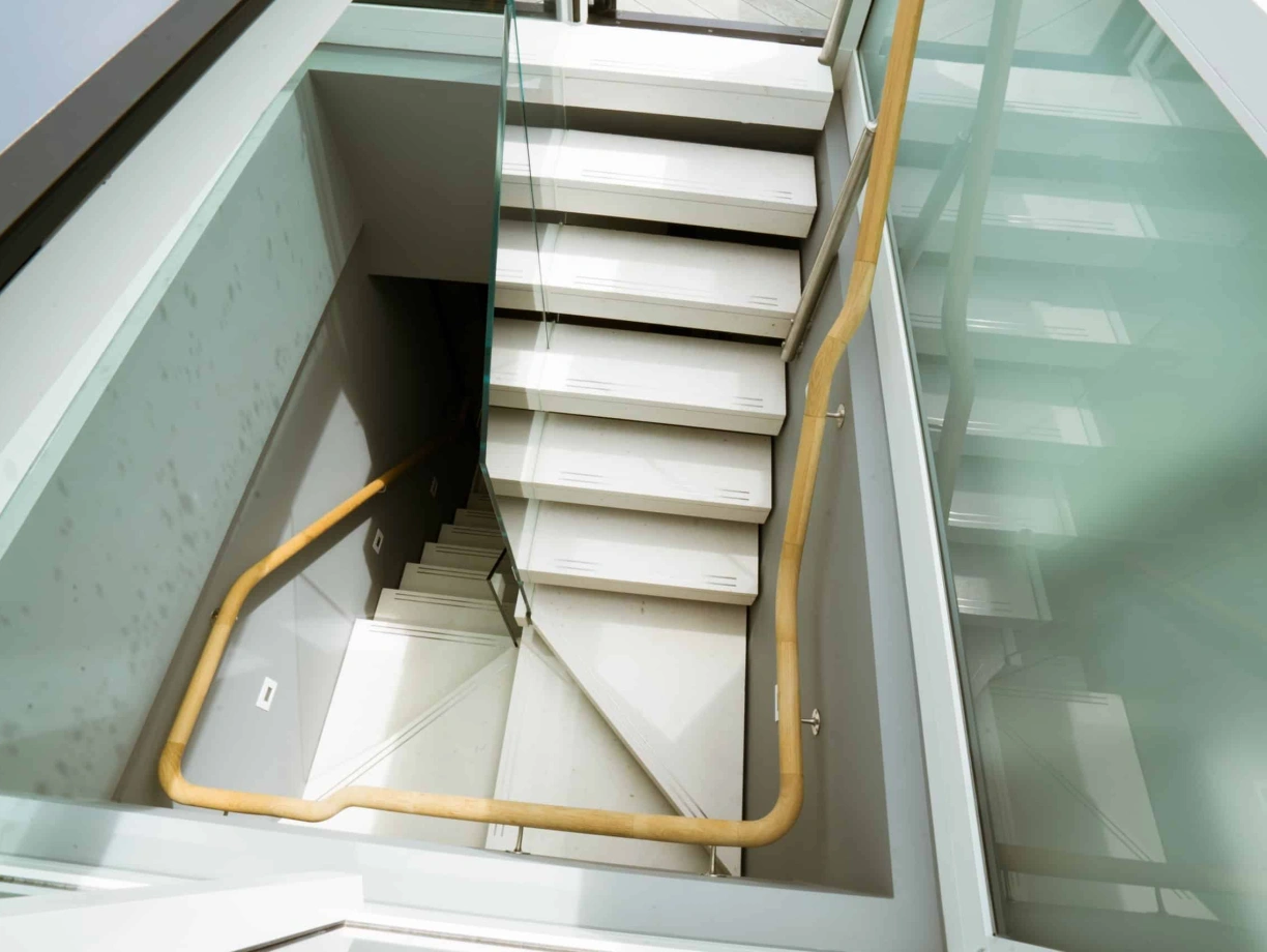 A modern staircase with wooden handrails and glass panels, leading down into a well-lit open space.