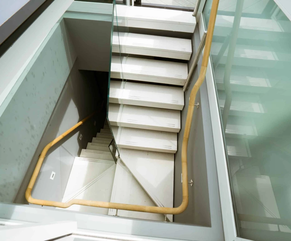 A modern staircase with wooden handrails and glass panels, leading down into a well-lit open space.
