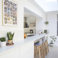 Cucina moderna con un banco bianco e seggioli di legno. Mostra una collezione di foto incorniciate su una parete. Piante verdi decorano il piano e l'illuminazione naturale entra da un lucernario.