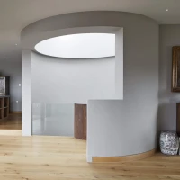 Bright and modern interior featuring a spacious open layout with wooden flooring, curved walls, and large windows. A dining area with colorful chairs is visible in the background, along with a dark wooden bookshelf. Elegant decor includes a vintage desk and a portrait on the wall.