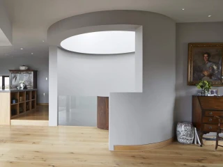 Bright and modern interior featuring a spacious open layout with wooden flooring, curved walls, and large windows. A dining area with colorful chairs is visible in the background, along with a dark wooden bookshelf. Elegant decor includes a vintage desk and a portrait on the wall.