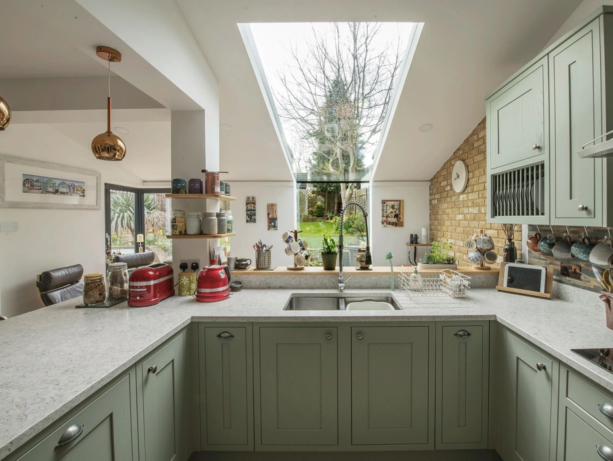 A modern kitchen features green cabinets, a white countertop, and a skylight that provides natural light. A view of a garden is visible through large windows, with various kitchen items and decor arranged on the counters.