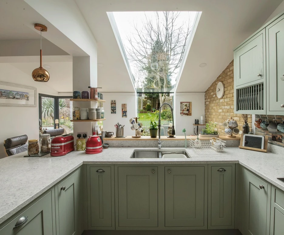 A modern kitchen features green cabinets, a white countertop, and a skylight that provides natural light. A view of a garden is visible through large windows, with various kitchen items and decor arranged on the counters.