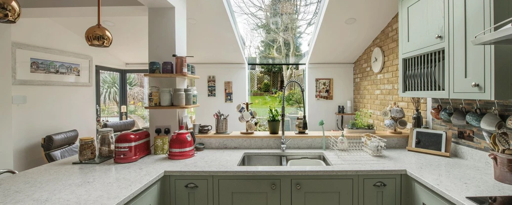 A modern kitchen features green cabinets, a white countertop, and a skylight that provides natural light. A view of a garden is visible through large windows, with various kitchen items and decor arranged on the counters.