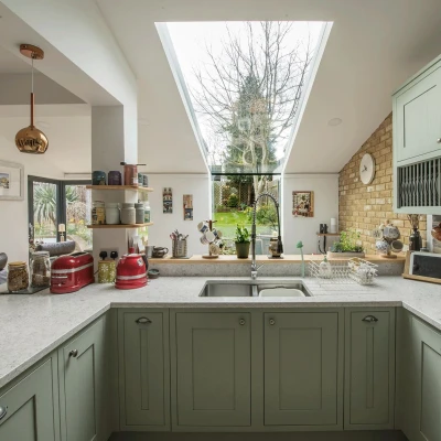 A modern kitchen features green cabinets, a white countertop, and a skylight that provides natural light. A view of a garden is visible through large windows, with various kitchen items and decor arranged on the counters.