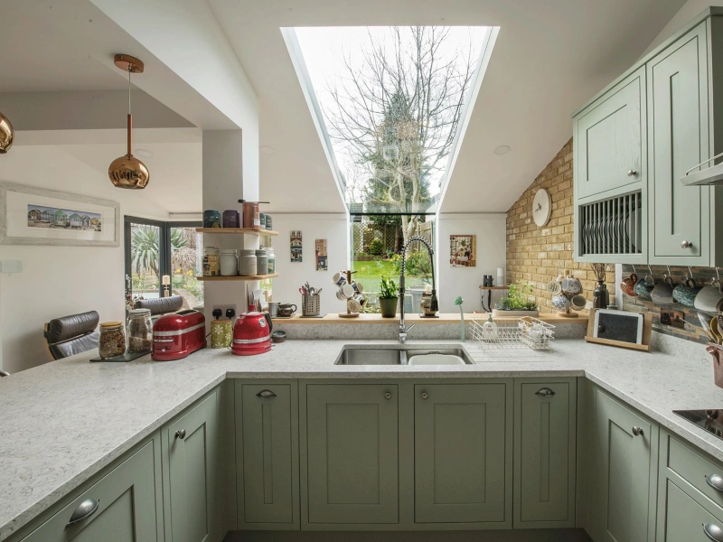 A modern kitchen features green cabinets, a white countertop, and a skylight that provides natural light. A view of a garden is visible through large windows, with various kitchen items and decor arranged on the counters.