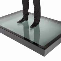 Eine Person steht auf einer großen Glasplatte, die auf einem dunklen Rahmen platziert ist. Der Schatten der Person ist auf der Glasoberfläche zu sehen. Die Schuhe der Person sind schwarz und sie trägt eine formelle Hose.