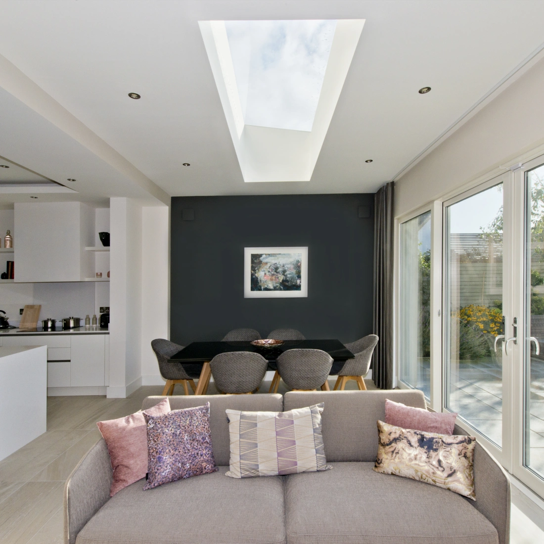 A modern living space with a light-colored sofa adorned with decorative pillows. The room features an open layout connecting to a dining area with a dark wall and a skylight above. Large glass doors lead to a garden filled with greenery.