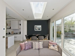 Sala moderna con un sofá en primer plano, cojines decorativos y un gran ventanal. Al fondo, una mesa de comedor rodeada por sillas. La luz natural entra a través de un tragaluz en el techo y se puede ver un jardín exterior.