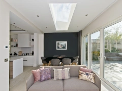 A modern living space with a light-colored sofa adorned with decorative pillows. The room features an open layout connecting to a dining area with a dark wall and a skylight above. Large glass doors lead to a garden filled with greenery.