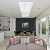 A modern living space with a light-colored sofa adorned with decorative pillows. The room features an open layout connecting to a dining area with a dark wall and a skylight above. Large glass doors lead to a garden filled with greenery.