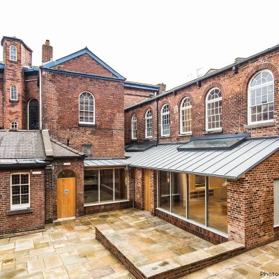 Brick buildings with large windows surround a paved courtyard. The architecture combines old and modern styles, featuring a glass extension. Overcast sky above.