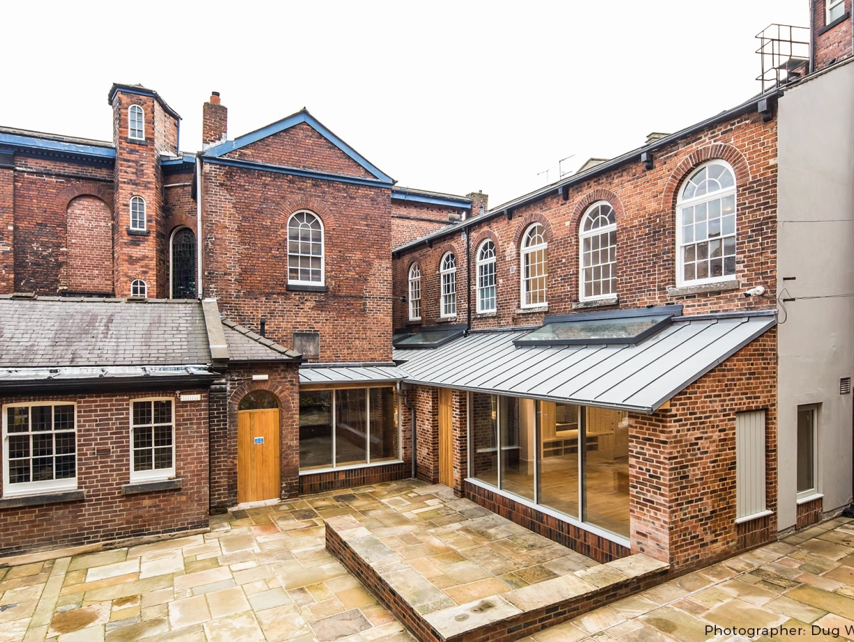 Brick buildings with large windows surround a paved courtyard. The architecture combines old and modern styles, featuring a glass extension. Overcast sky above.