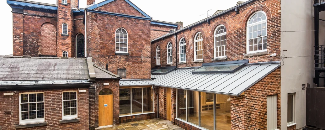 Brick buildings with large windows surround a paved courtyard. The architecture combines old and modern styles, featuring a glass extension. Overcast sky above.