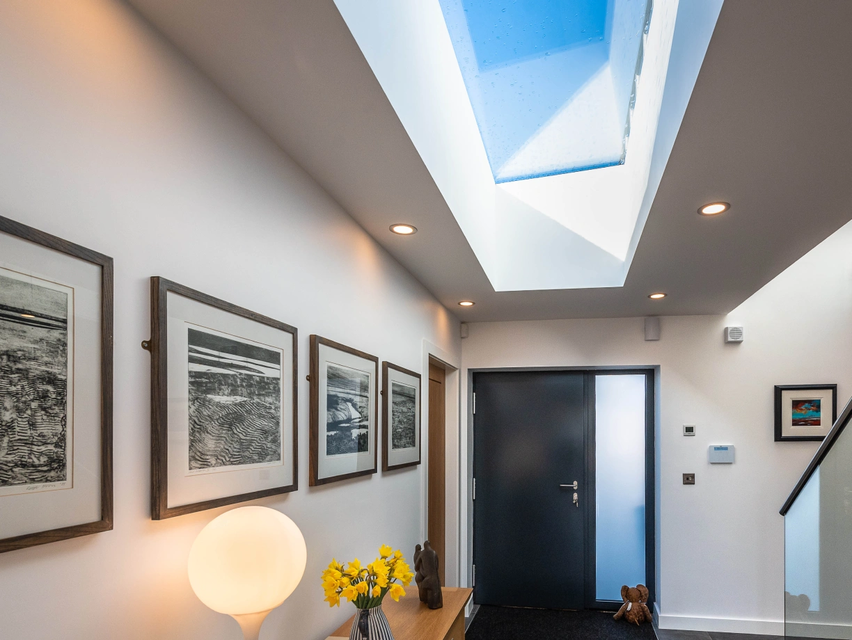 A modern hallway featuring a sleek wooden console table with a lamp and a vase of yellow flowers. Framed black-and-white artwork hangs on the walls, and a large skylight allows natural light to illuminate the space. A dark door leads outside, and a small rug is placed at the entrance.