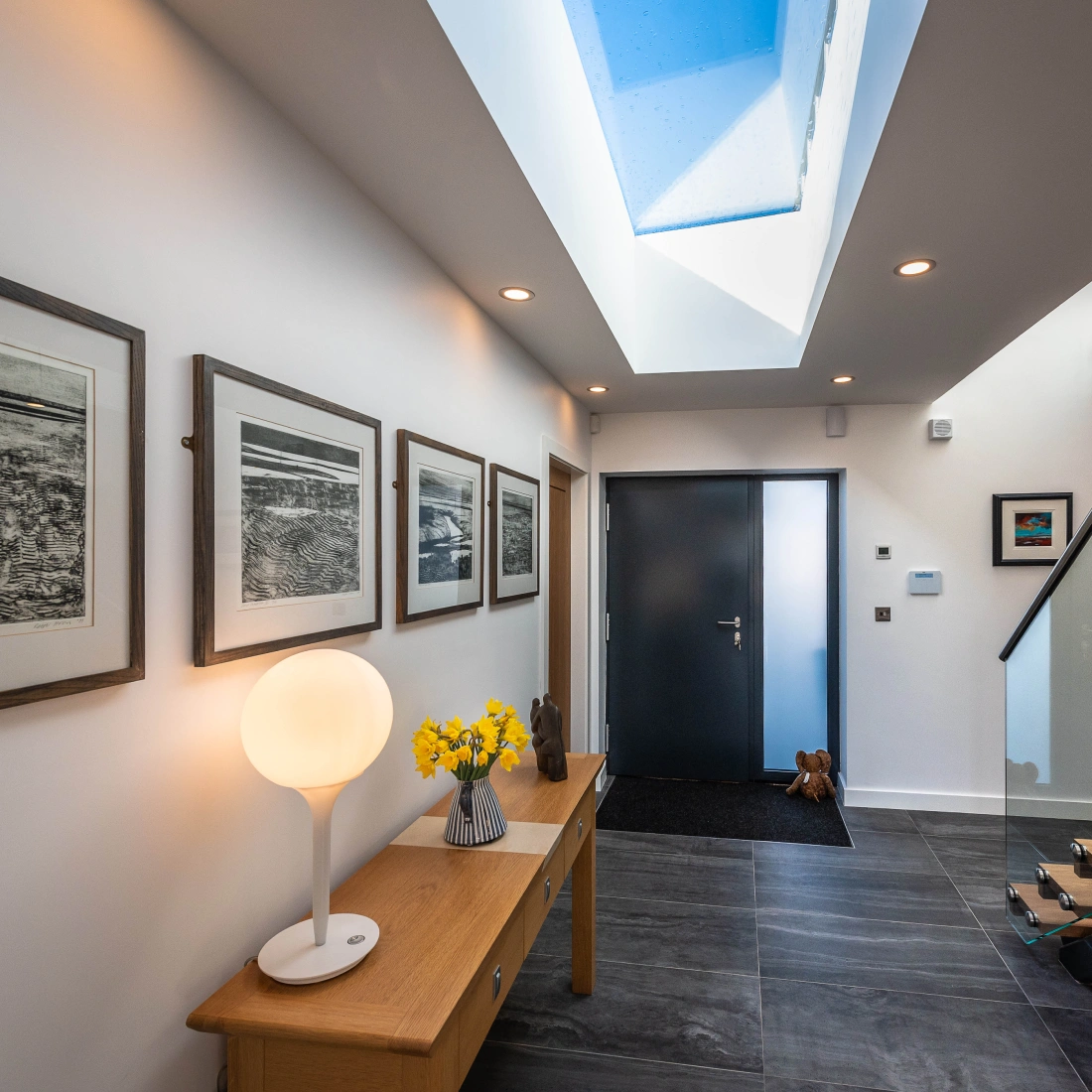 A modern hallway features a dark front door and a light wood console table with a round lamp and a vase of yellow flowers. Walls are adorned with framed artwork, and a skylight above lets in natural light. Decorative elements include a small sculpture and a rug at the entrance.