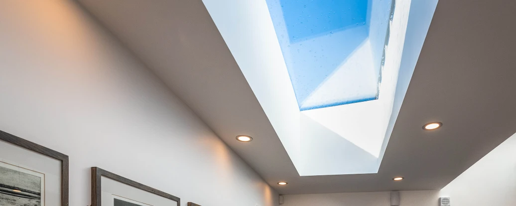A modern hallway featuring a sleek wooden console table with a lamp and a vase of yellow flowers. Framed black-and-white artwork hangs on the walls, and a large skylight allows natural light to illuminate the space. A dark door leads outside, and a small rug is placed at the entrance.