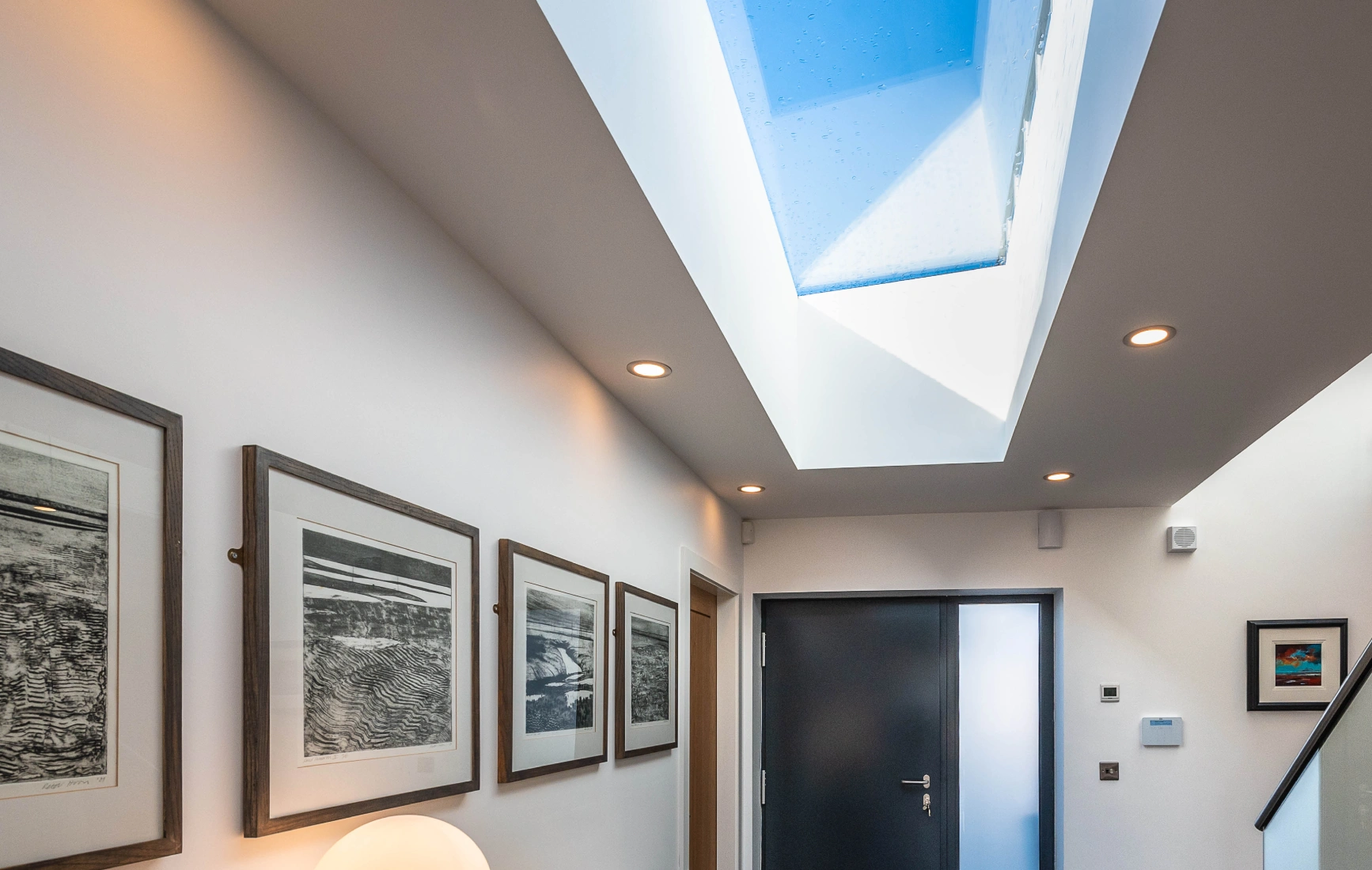 A modern hallway featuring a sleek wooden console table with a lamp and a vase of yellow flowers. Framed black-and-white artwork hangs on the walls, and a large skylight allows natural light to illuminate the space. A dark door leads outside, and a small rug is placed at the entrance.