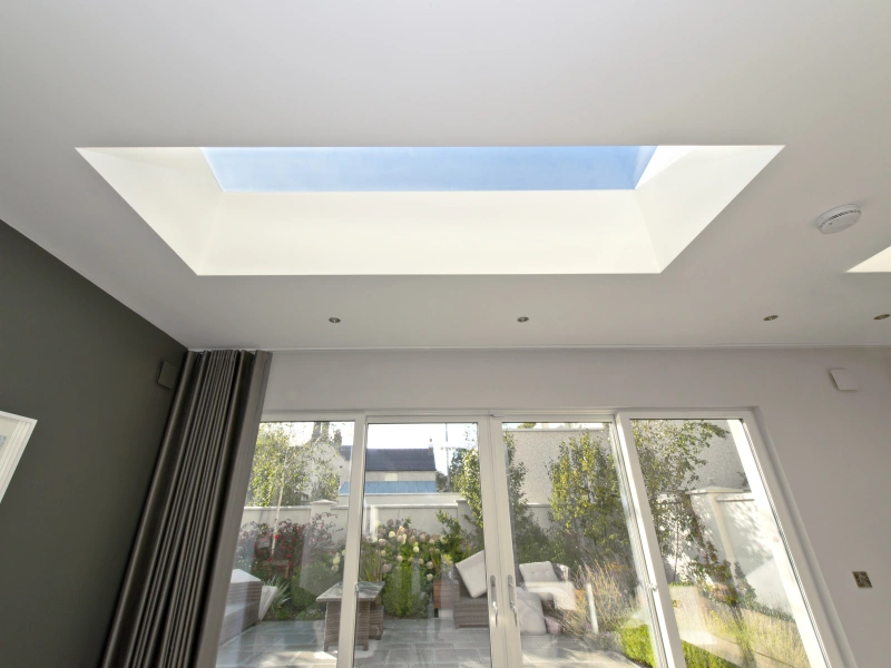 Bright interior space featuring a skylight that frames the blue sky. Large glass doors lead to a garden with greenery and outdoor seating. Walls have a modern design with a dark accent wall and light-colored ceiling.