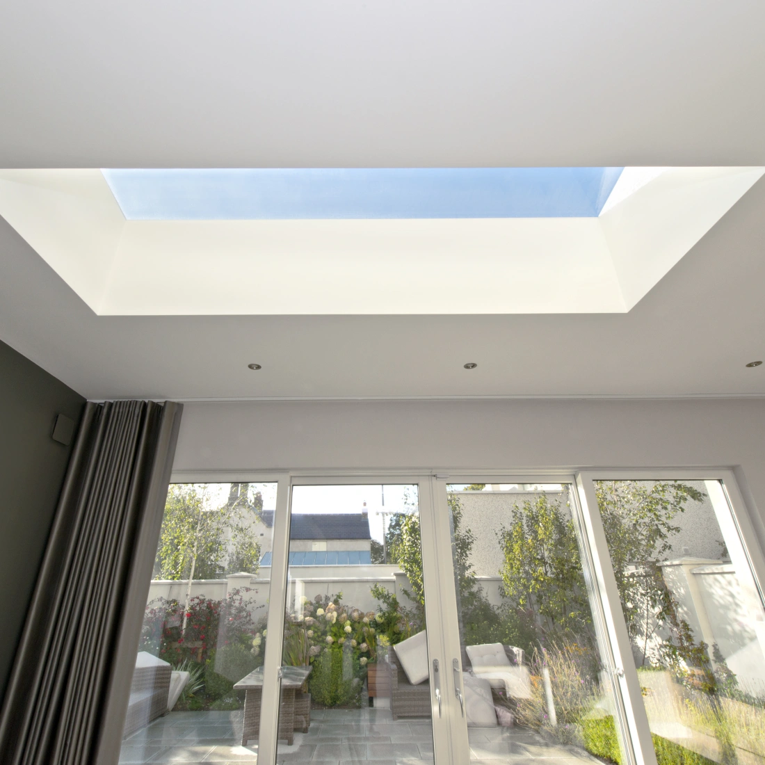 Bright interior space featuring a skylight that frames the blue sky. Large glass doors lead to a garden with greenery and outdoor seating. Walls have a modern design with a dark accent wall and light-colored ceiling.