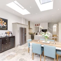A modern kitchen featuring a long wooden dining table with blue chairs, a large fridge, and a stylish side cabinet. Skylights illuminate the space, and a vibrant painting hangs on the wall. A sign reading "EAT" decorates the room, which has a mix of contemporary and rustic design elements.