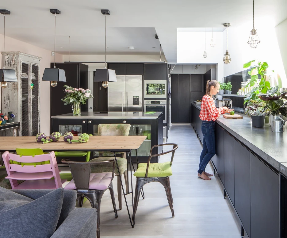 A modern kitchen featuring black cabinets and a large skylight. A woman in a checkered shirt stands at a sleek counter, preparing food. The dining area includes a rustic table surrounded by colorful chairs, with plants and flowers adding decor throughout the space.