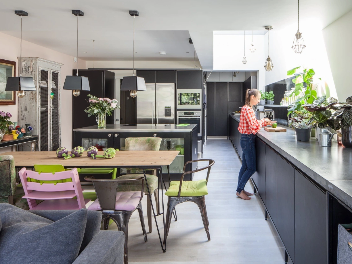 A modern kitchen featuring black cabinets and a large skylight. A woman in a checkered shirt stands at a sleek counter, preparing food. The dining area includes a rustic table surrounded by colorful chairs, with plants and flowers adding decor throughout the space.