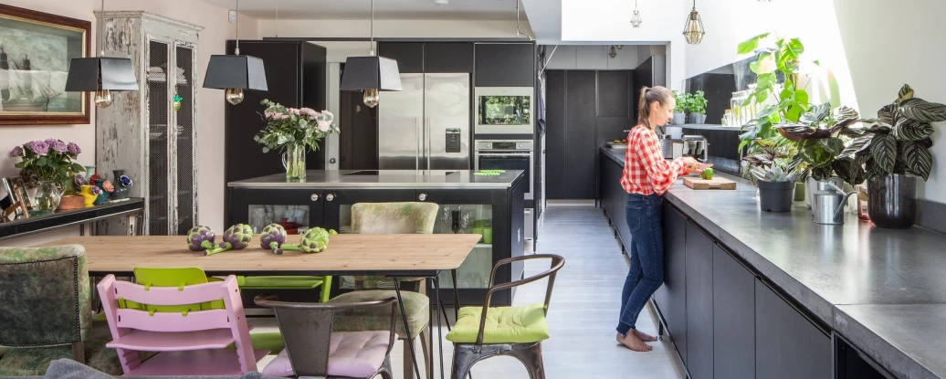 A modern kitchen featuring black cabinets and a large skylight. A woman in a checkered shirt stands at a sleek counter, preparing food. The dining area includes a rustic table surrounded by colorful chairs, with plants and flowers adding decor throughout the space.