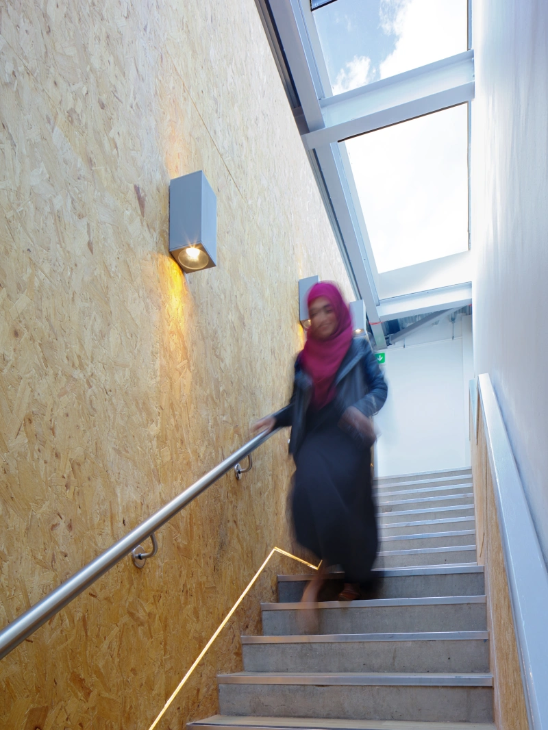 A woman wearing a pink hijab and a long black dress descends a set of modern stairs. The stairs are complemented by warm lighting and a wooden wall. Natural light filters in through a glass window above.
