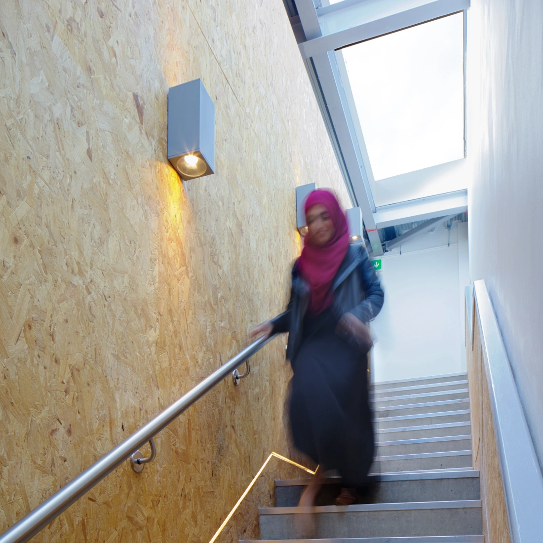 A woman wearing a pink hijab and a long black dress descends a set of modern stairs. The stairs are complemented by warm lighting and a wooden wall. Natural light filters in through a glass window above.