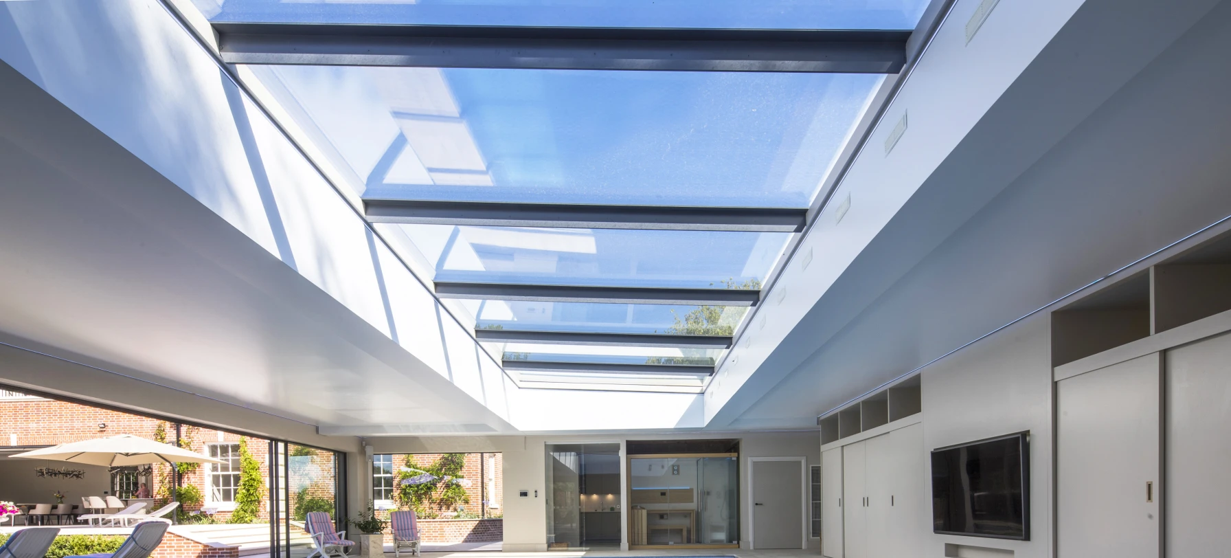 A spacious indoor pool area with a bright skylight, featuring a clear blue pool and modern minimalist design. Sliding doors lead to an outdoor space with lounge chairs.