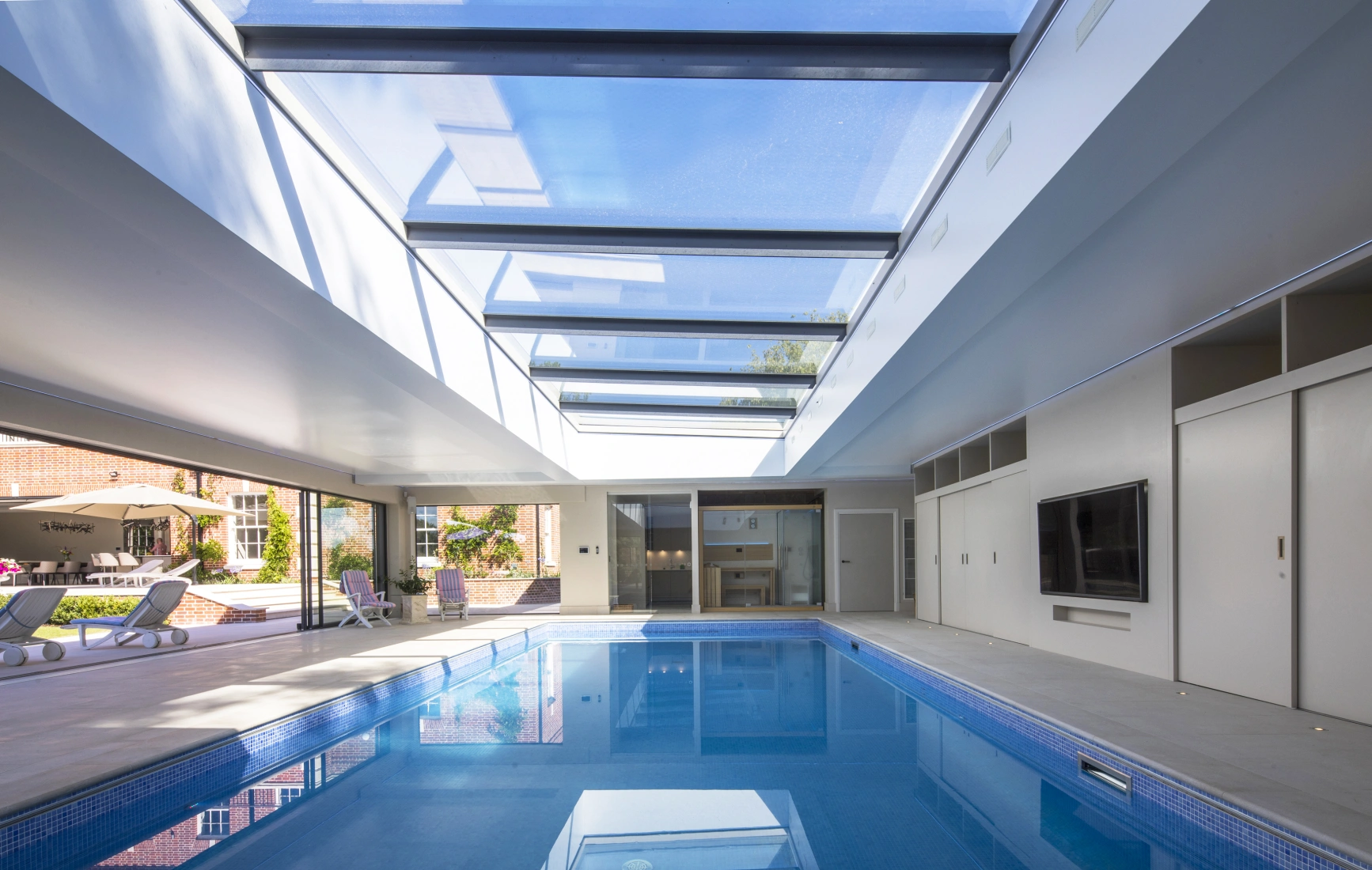 Interior de una casa con piscina iluminada por un techo de vidrio. Las paredes son de color claro y hay un área de descanso con sillas. Se puede ver el exterior con un jardín al fondo.